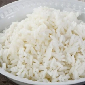 Eine Nahaufnahme eines weißen Reisgerichts in einer weißen Schüssel auf einem Holztisch. Der Reis ist fluffig und leicht dampfend.