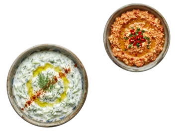 Draufsicht auf eine kleine Tonschale mit cremigem, hausgemachtem Tzatziki und eine danebenstehende Tonschale mit würziger Tirokafteri-Käsecreme, isoliert auf einem neutralen Hintergrund